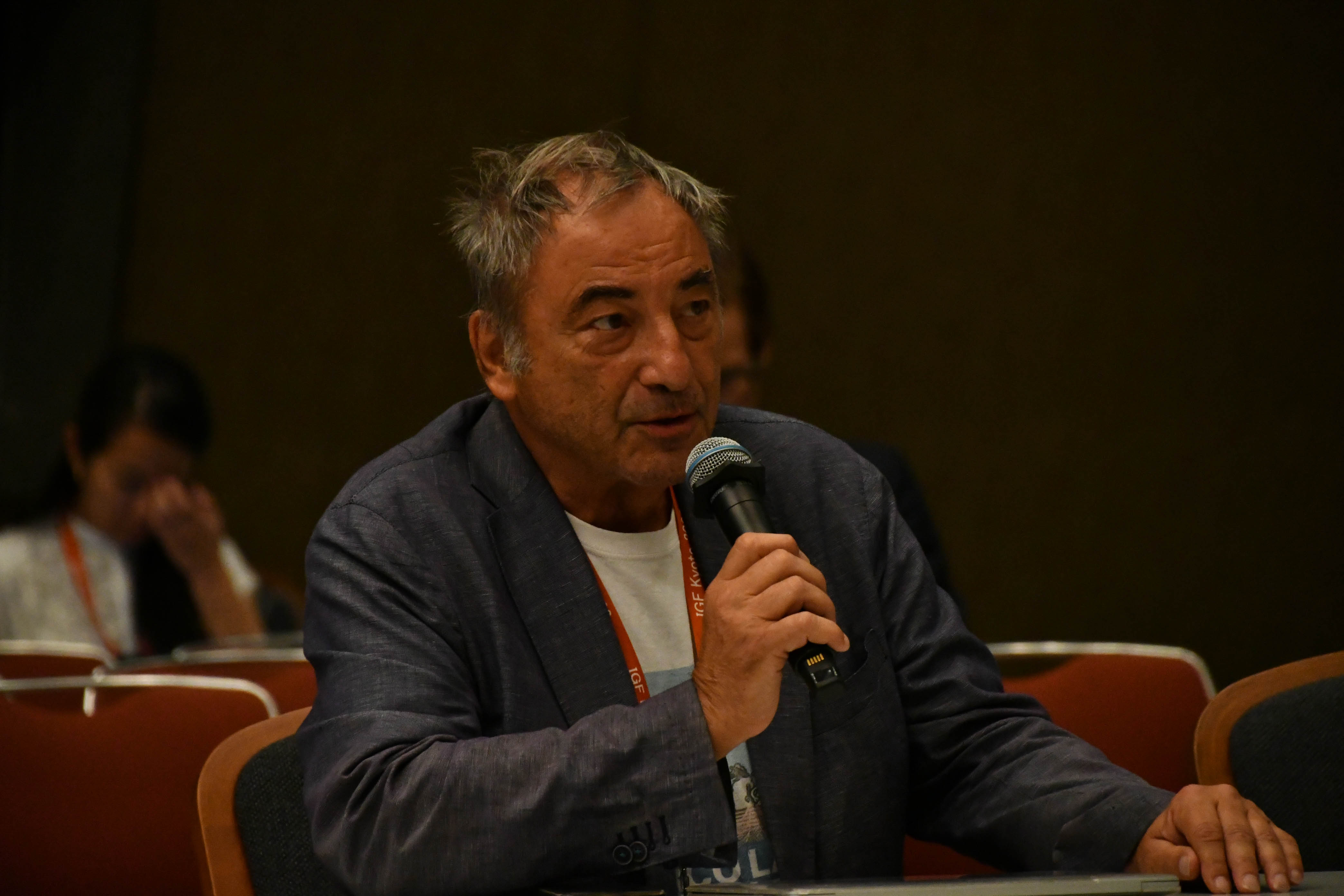 Man with a dark blue jacket seated at a table and speaking on a microphone